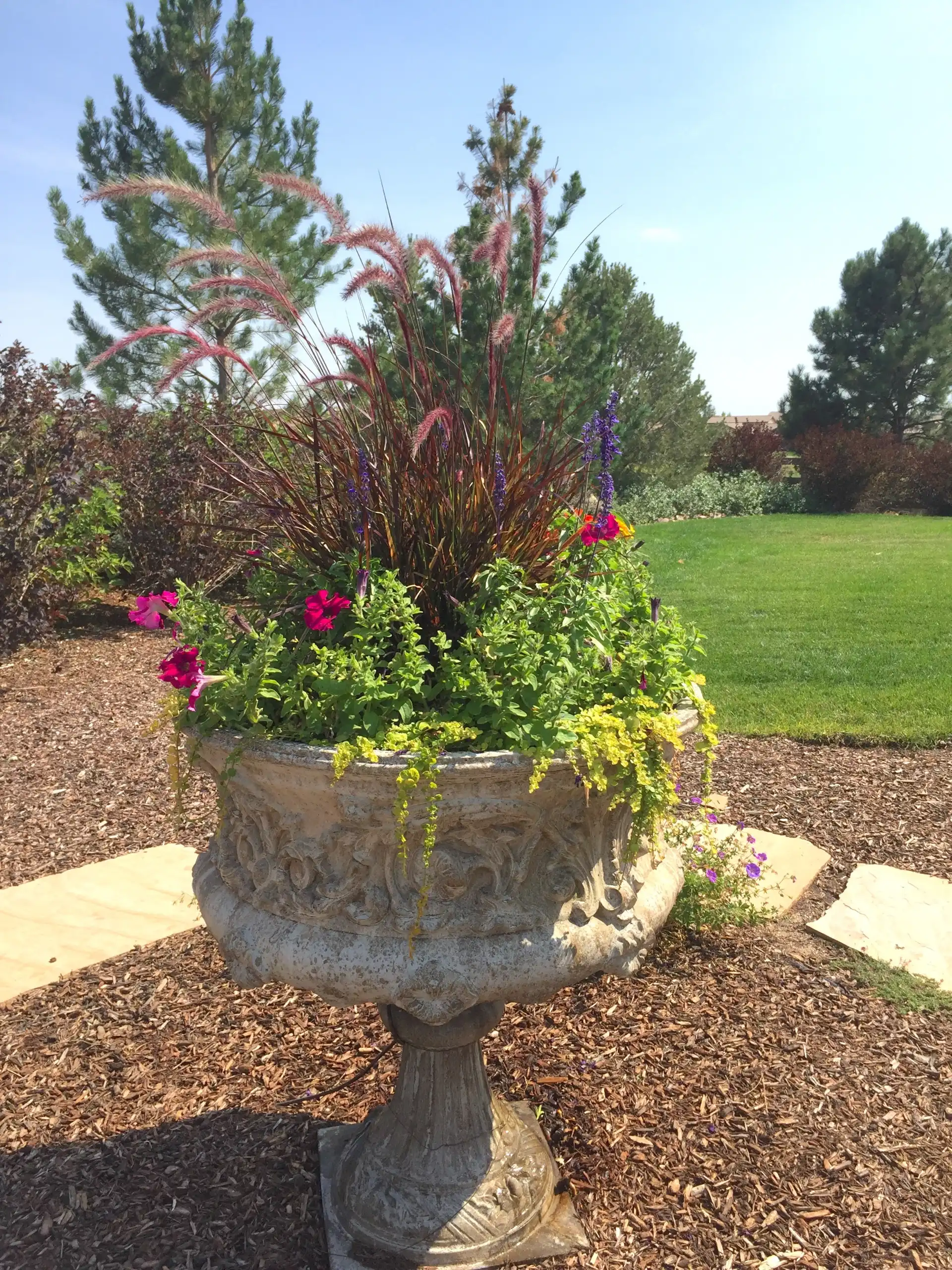 Ornate stone urn planter filled with purple fountain grass and bright flowers.