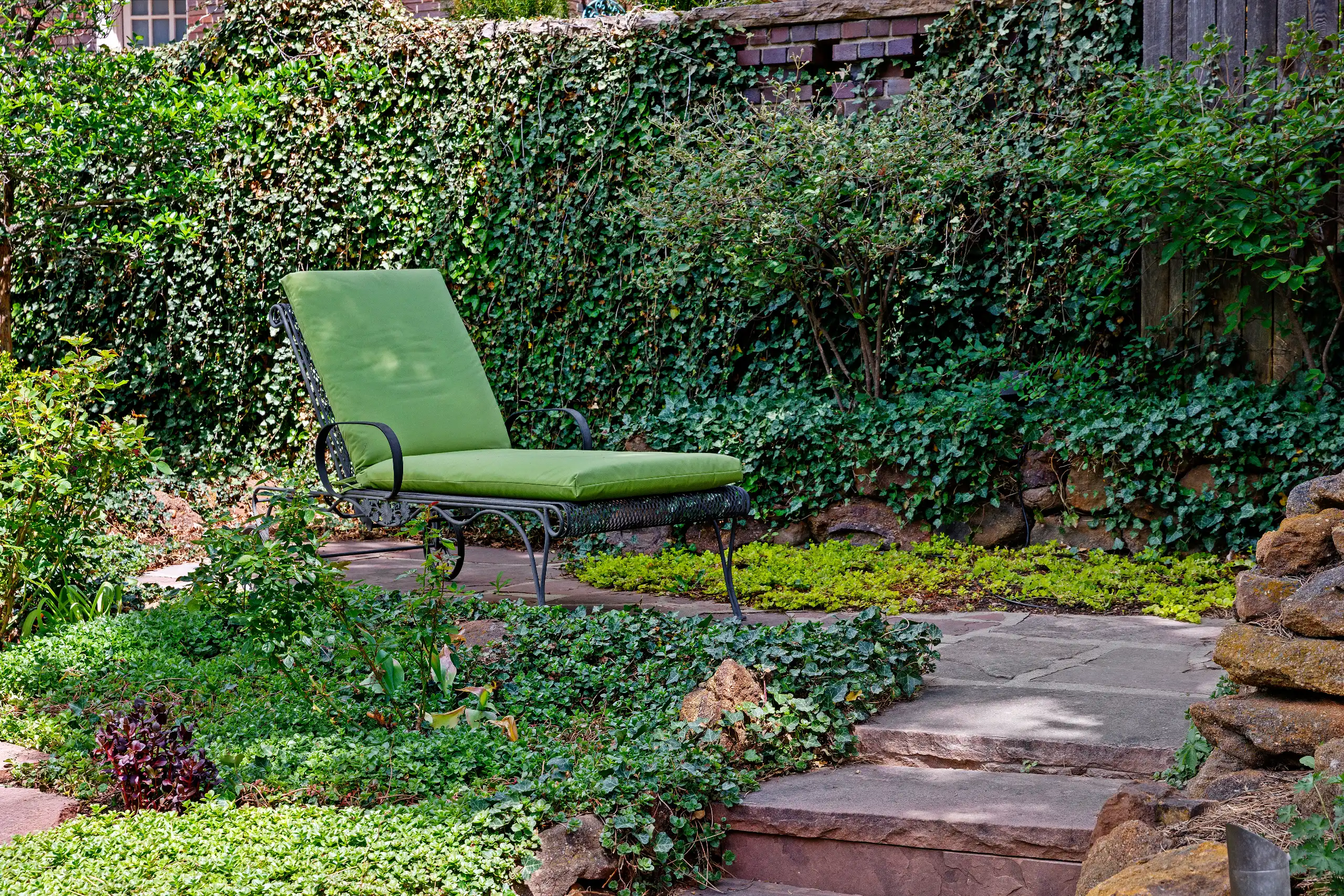 Green lounge chair on a stone patio surrounded by ivy and lush garden greenery.